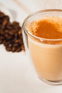 A warm latte in a glass cup topped with cinnamon, surrounded by coffee beans.