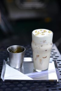 Delicious iced latte topped with nuts served on a metal tray, accompanied by a metal cup.