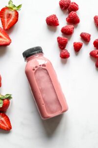 Top view of fresh strawberry and raspberry smoothie in a bottle with scattered fruits on white background.
