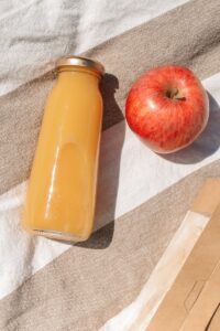 A bottle of apple juice and a fresh apple on a striped picnic blanket outdoors.