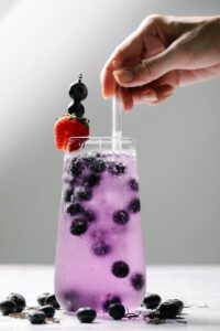 Close-up of a hand holding a glass of sparkling blueberry drink with a strawberry garnish.